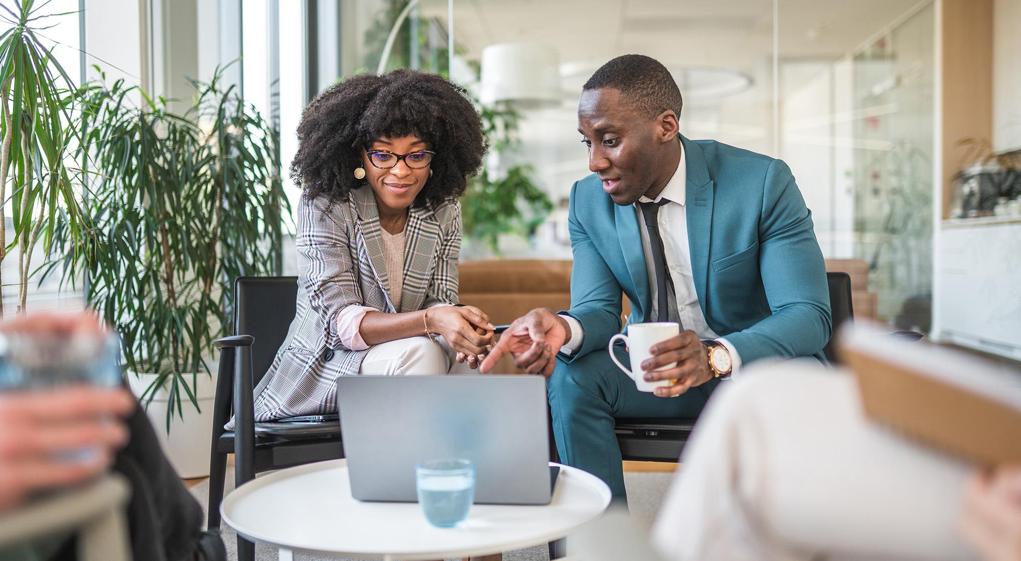 woman and man in business setting seated on couch and talking while looking at laptop