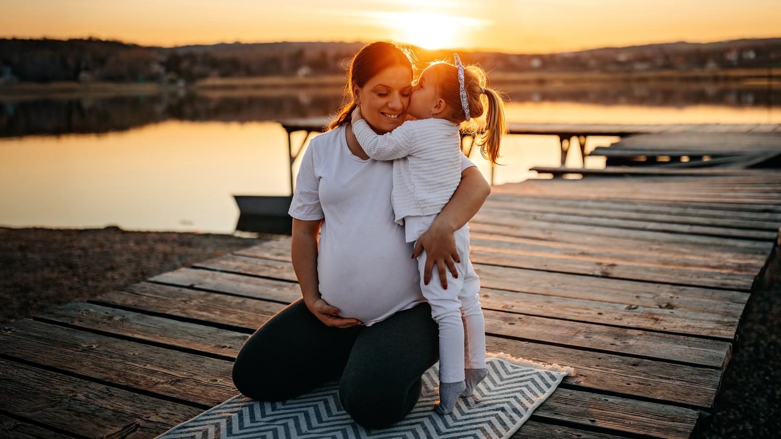 Pregnant woman hugging her young daughter near a lake.