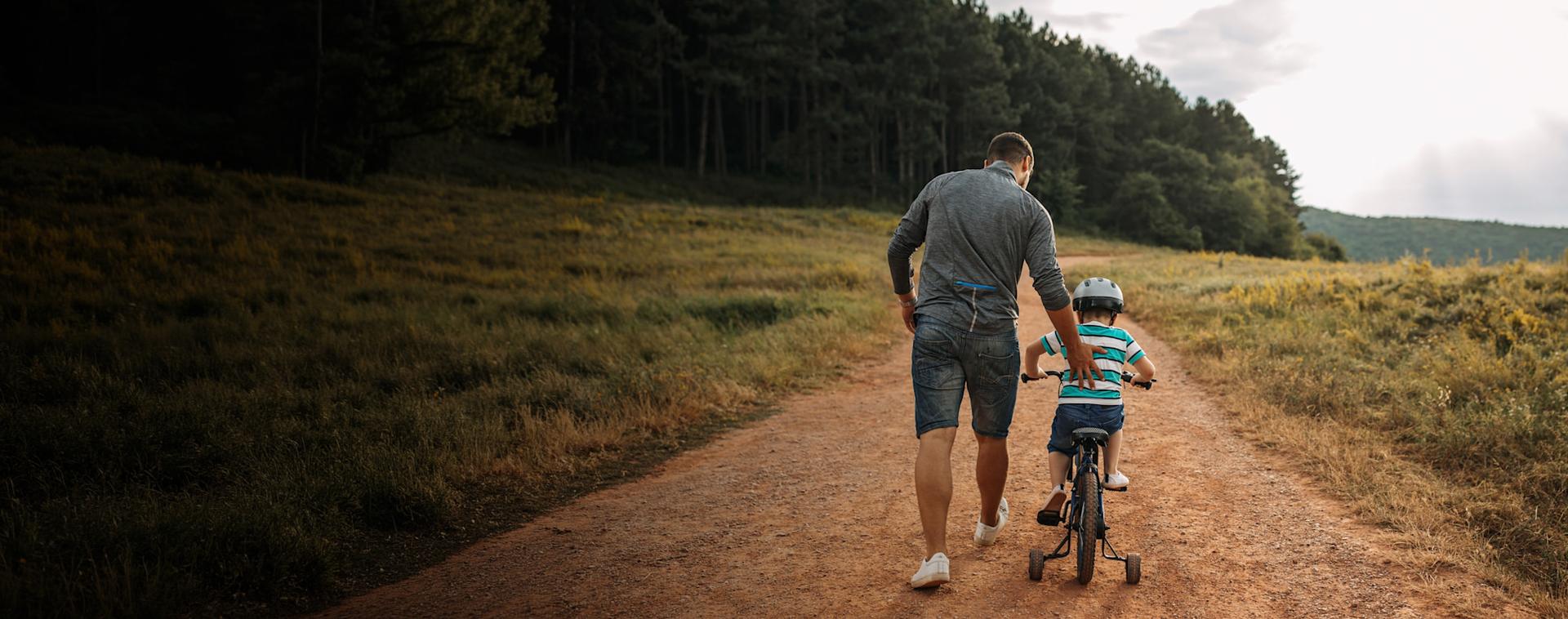 AR25 Hero image A father helps his son ride a bike down a path in a field.