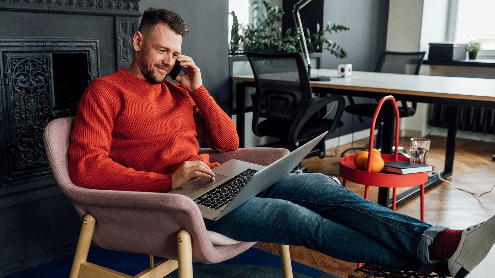 Man-talks-on-phone-while-looking-at-laptop-seated-in-chair