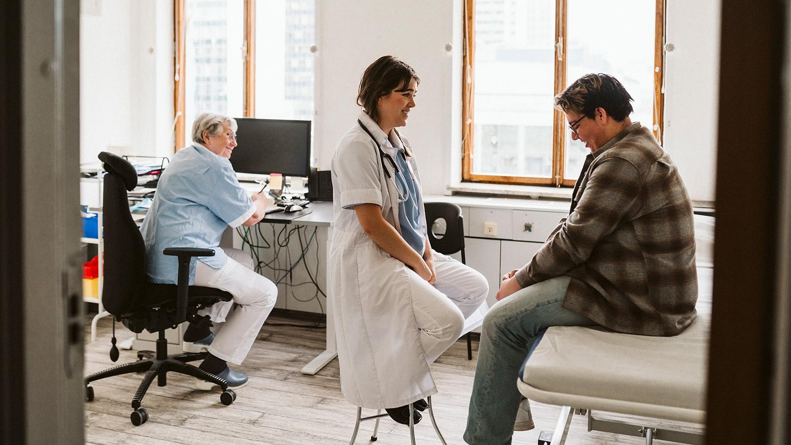 Patient in doctor's office with head down but smiling doctor showing concern another women seated at computer