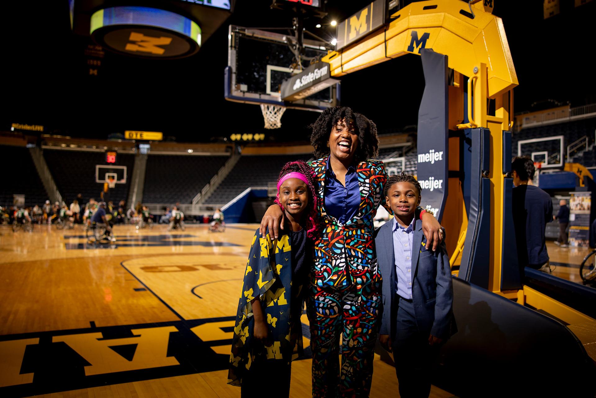 A woman in a colorful suit stands on a basketball court with her young twins, a boy and a girl.
