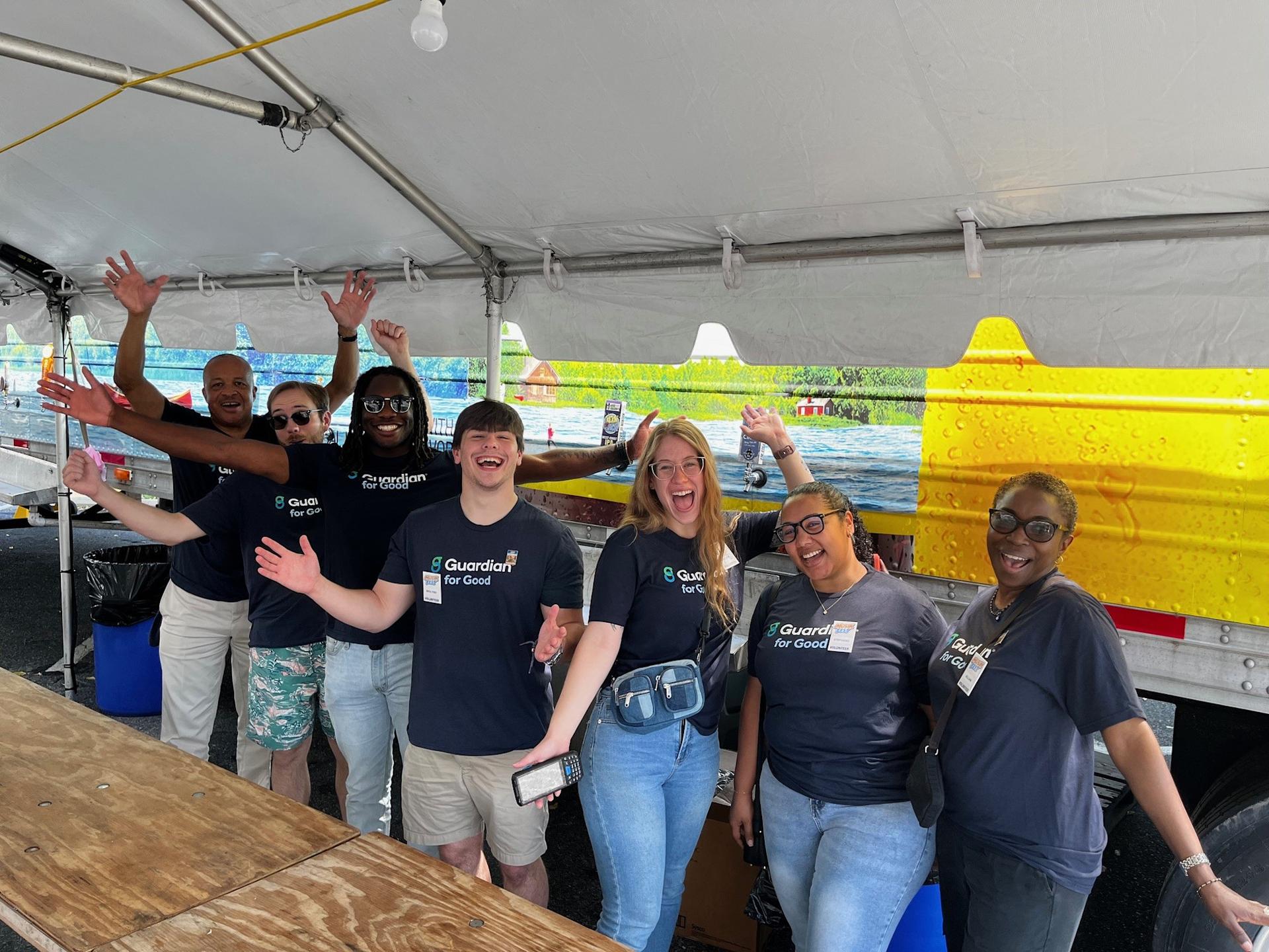 A group of Guardian for Good volunteers standing under an event tent, wearing matching Guardian for Good T‑shirts and raising their arms toward the camera.