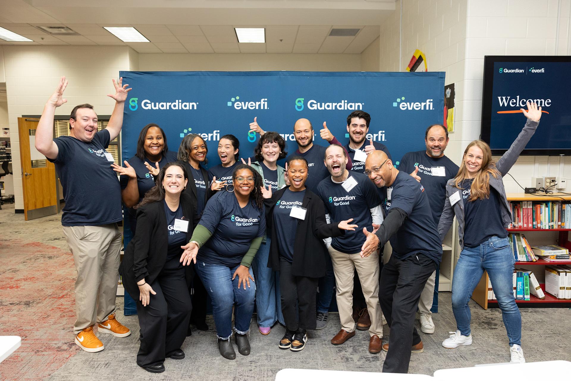 A group of colleagues in matching shirts smile and give a thumb's up in front of a step and repeat.