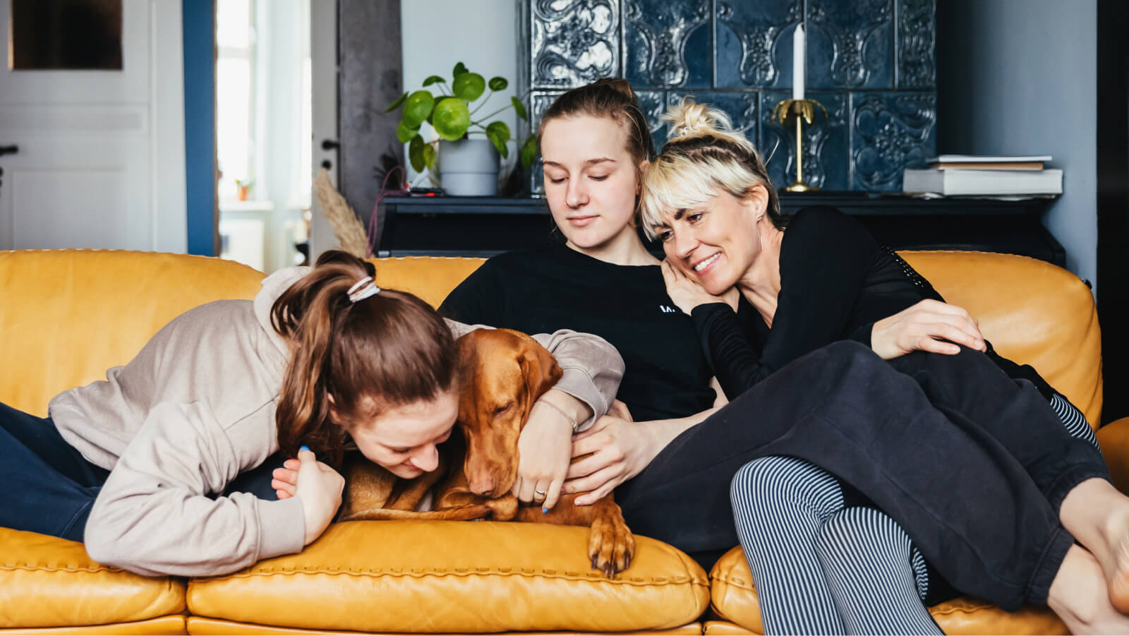 Mother happily relaxing on couch and spending time with teenage daughters and dog 