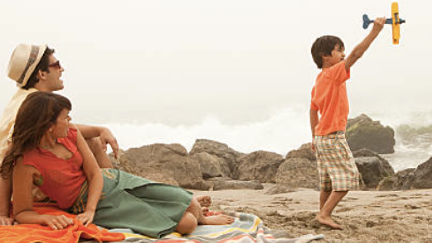 Family at the beach. The parents are watching their little body play with a toy airplane.