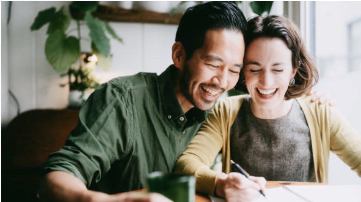 Smiling couple sitting at a table looking at a page in a notebook. 