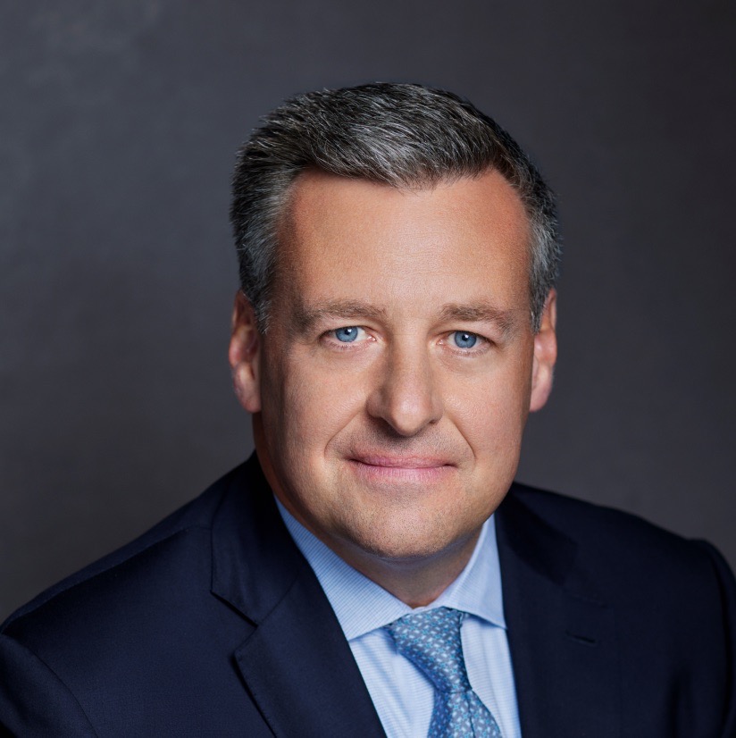 Andrew J McMahon looks at the viewer confidently, wearing a dark blue suit, crisp button-up shirt, and a blue tie with white spots. His thick grey hair is combed to the right.