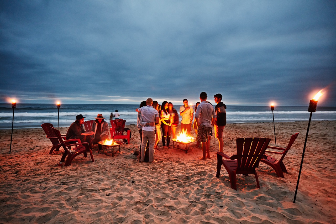 A group of twelve friends in their 20's stand on the beach at dusk, circled around an outdoor fire pit. Behind them, dark blue ocean waves lap onto the shore. Some sit in red wooden beach chairs.