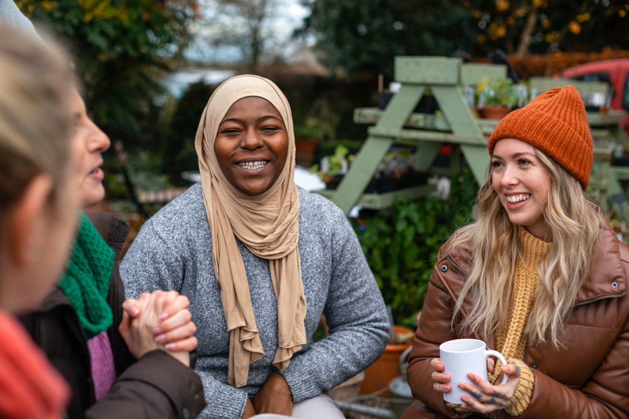 Two friends sit around a picnic table, speaking with a third friend whose hands peek into the frame of the photo. Both friends are smiling brightly, while wearing warm, outdoor clothing.