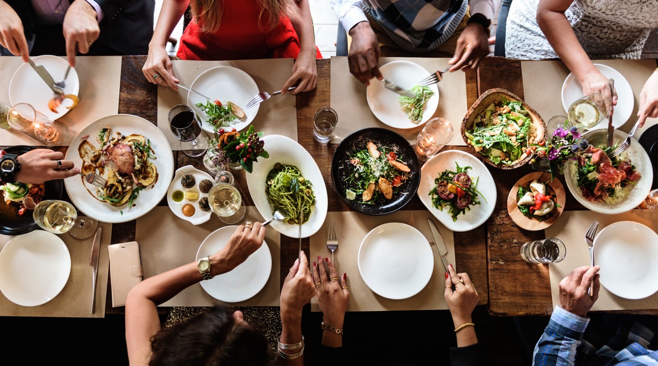 An overhead shot of a reclaimed wooden table, set for dinner. It's filled with lush salads, vegetables and green pastas. Eight friends reach for food from large shared plates, or eat off their own small plates. We see none of their faces, just their arms and hands. Some wear beautiful rings, pearl bracelets or large watches. It's clear they're dressed for a special occasion.