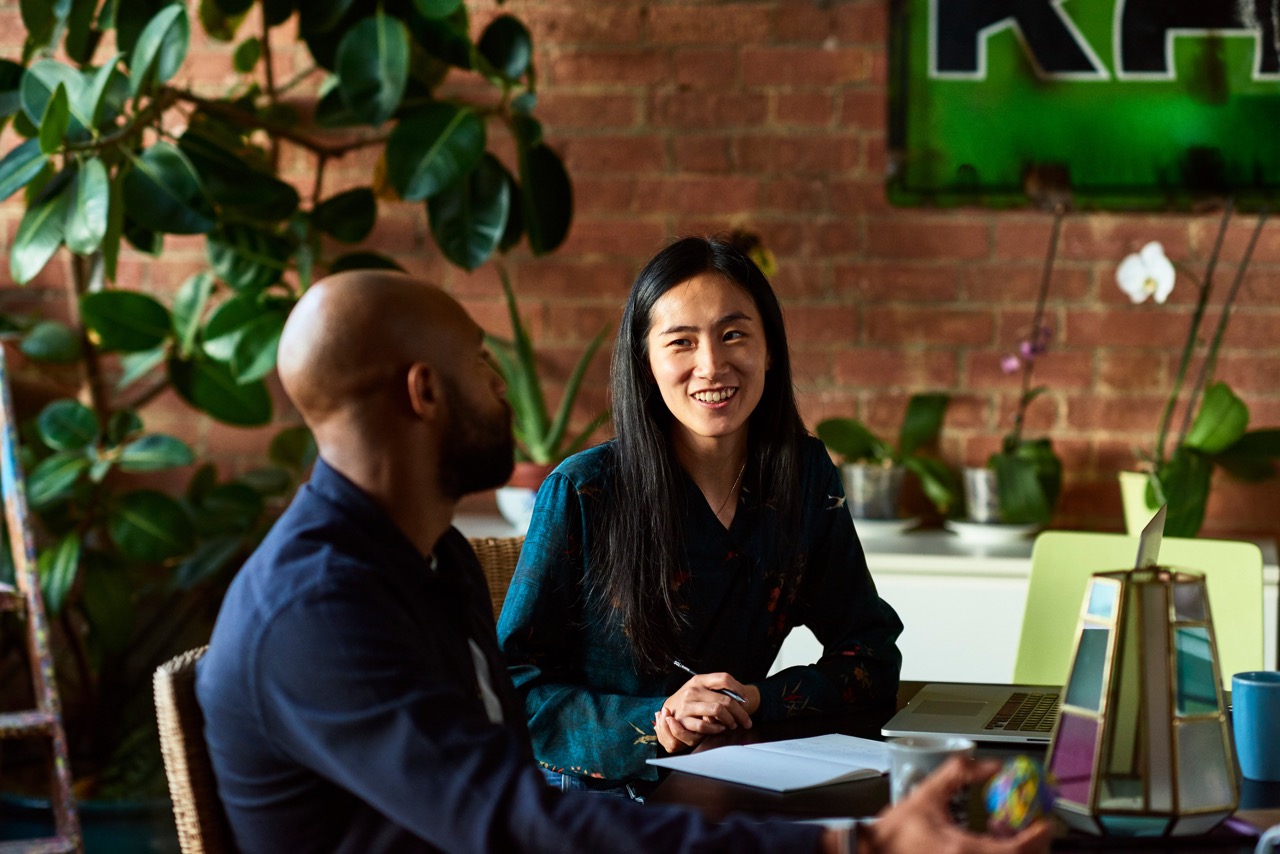 Two colleagues sit around a small coffee table. One holds a pen, sitting in front of a small amount of paperwork. They smile at each other.