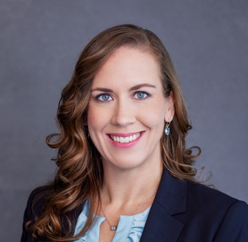 Erin Culeck looks straight into the lens, her mouth and eyes smiling wide. She wears a blue blazer with a blue shirt, and her hair is curled shoulder length. She wears small, tasteful earrings with a matching necklace.