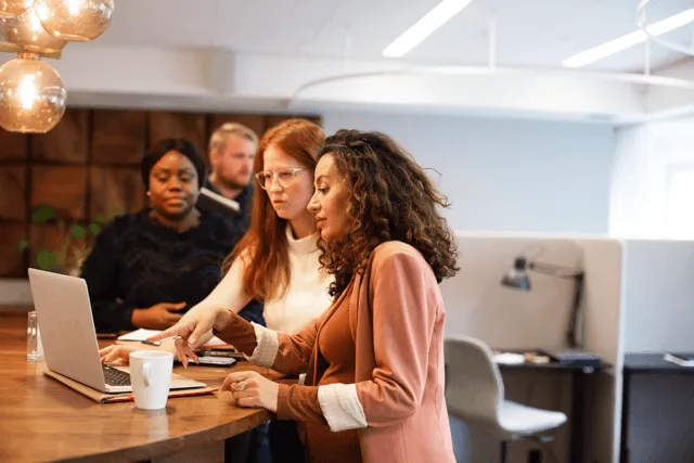 Three multi-ethnic women standing at a desk, looking over a laptop.