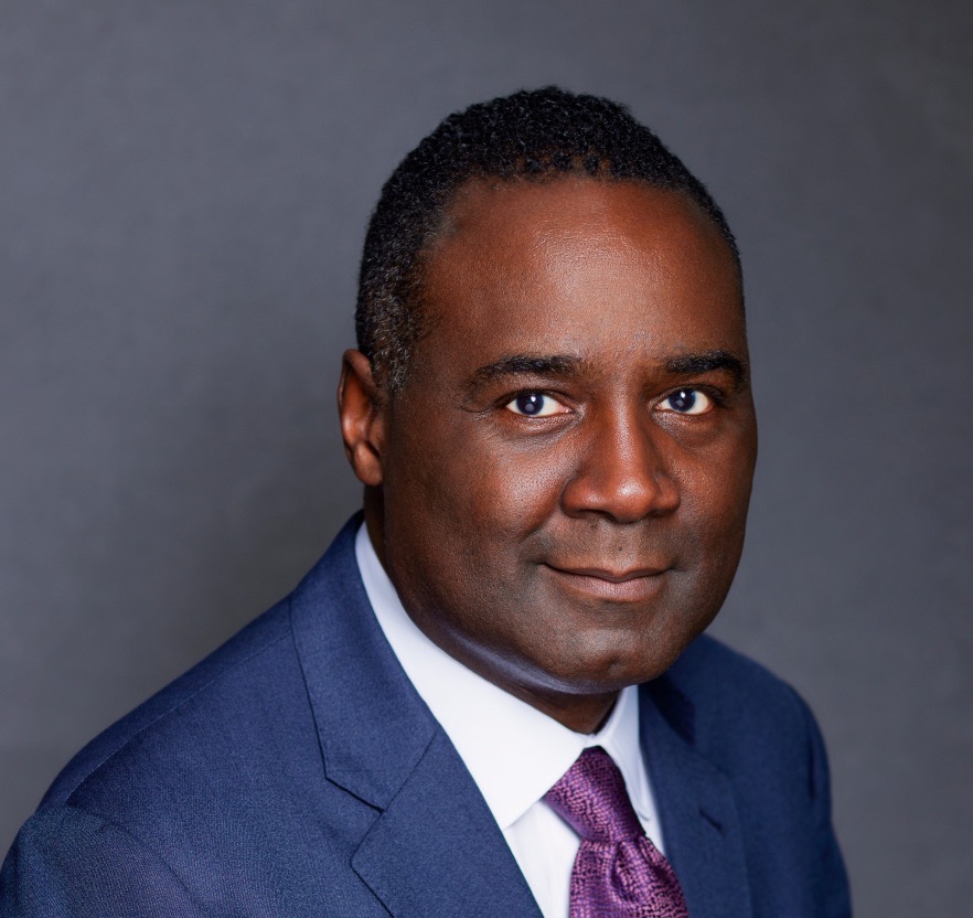 Kermitt Brooks looks at the camera with a small smile, his body angled to the right. He wears a blue suit, a crisp white dress shirt, and a purple patterned tie. He has dark, close-cropped hair.