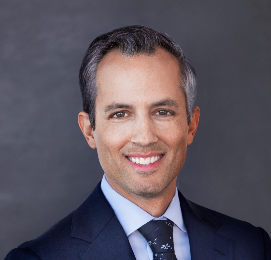 Kevin Molloy smiles broadly into the camera, wearing a dark blue suit, light blue dress shirt, and a blue tie with small white flowers on it. His short hair greys at the temples.