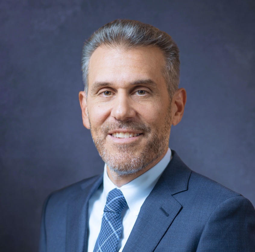 Nick Liolis looks warmly into the camera, wearing a light blue suit jacket, white shirt and blue tie with a light criss-cross pattern. He has a very short beard and short salt-and-pepper hair.