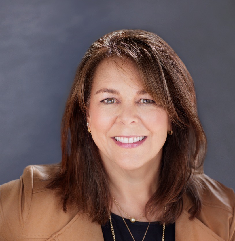 Stacey Hoin looks powerfully into the camera, wearing a tan leather jacket with a black shirt underneath. She wears three thin gold necklaces, and has shoulder length brown hair. 