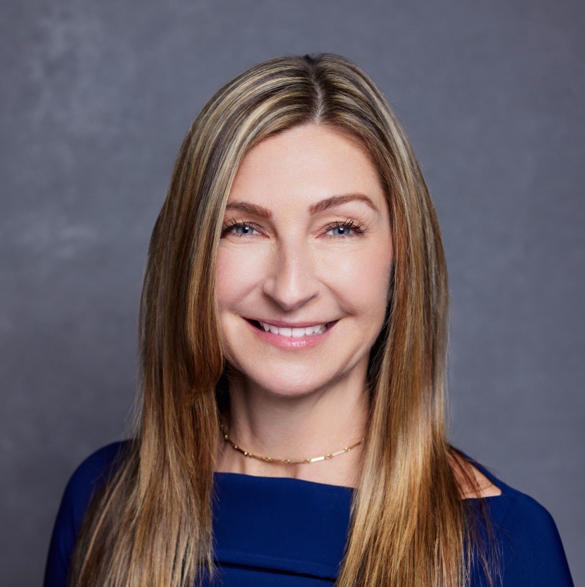 Wendy Wahl smiles widely into the camera, wearing a dark blue dress. Her shoulder length hair is streaked with blonde, and she wears a thin gold chain around her neck.